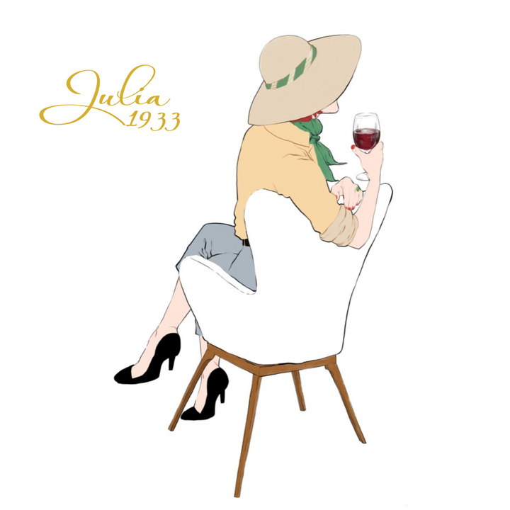 Illustration of a woman with a wide-brimmed hat, green scarf, beige top, and gray pants on a white chair holding wine. Julia 1933 Hand Soap &amp; Cream Duo by Hummingbird Farms sits nearby. "Julia 1933" is written elegantly to the left.
