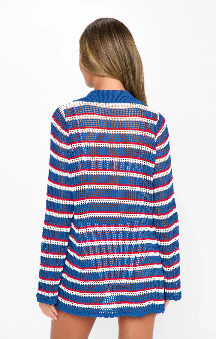 A woman with long hair stands facing away, wearing the Zertz BUTTON UP COVERUP—a long-sleeve, open-knit striped sweater in blue, red, and white—perfect for your summer wardrobe. The background is plain white.