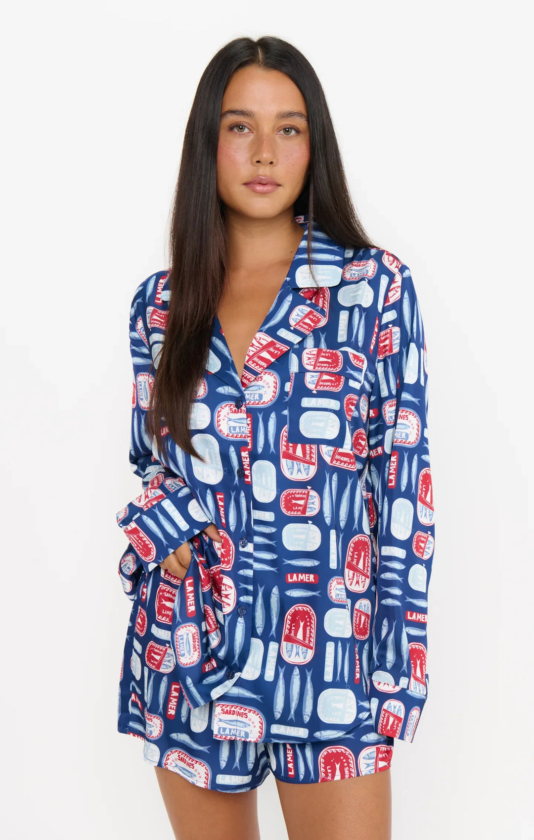 A woman with long dark hair wears the Zertz SARDINE BEAUTY SLEEP PJ SET, featuring a playful blue, red, and white Salty Sardines print. The silky button-up top and matching shorts have a relaxed fit. She poses against a plain white background with one hand in her pocket.