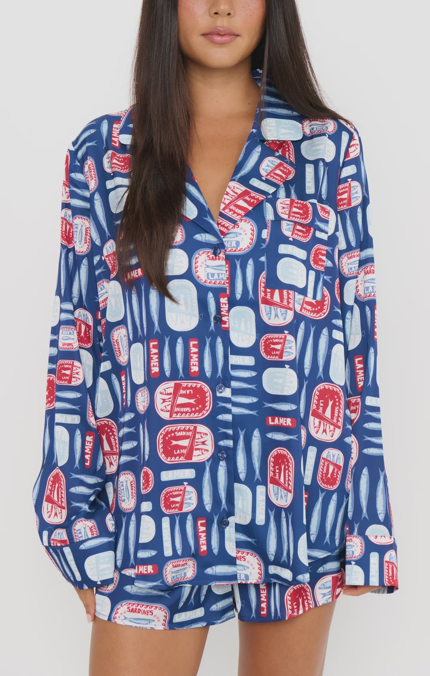 Wearing Zertz’s SARDINE BEAUTY SLEEP PJ SET in the Salty Sardines print—a blue base with red and white fish cans, LAMER text, and sardine tins—a relaxed button-up top pairs perfectly with comfy, loose-fitting shorts.