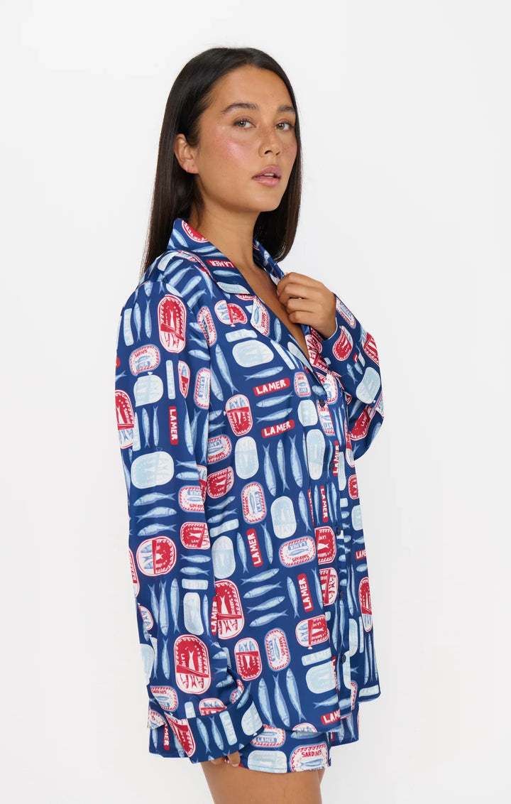 A woman with long dark hair models the Zertz SARDINE BEAUTY SLEEP PJ SET, featuring a relaxed button-up top in a blue and red Salty Sardines print, standing against a plain white background and gazing to the side.