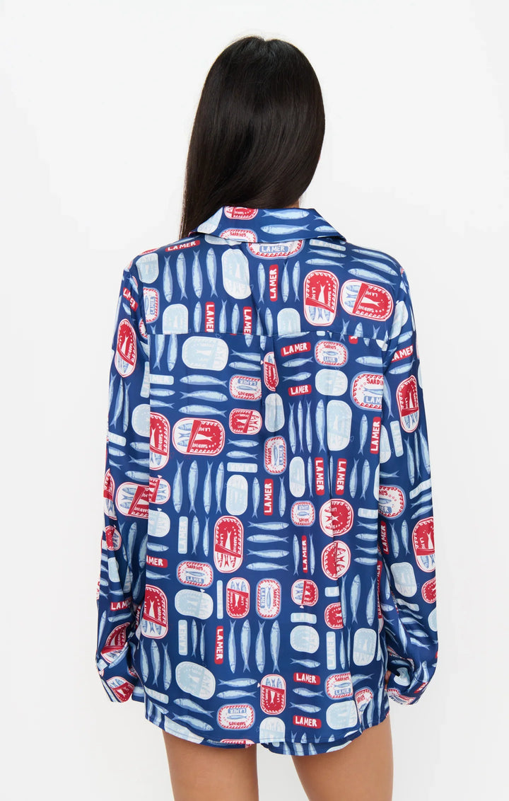 A person with long dark hair stands facing away, wearing the Zertz SARDINE BEAUTY SLEEP PJ SET—a relaxed blue button-up top with red and white rectangles, ovals, and playful Salty Sardines graphics, plus the word "Laker.