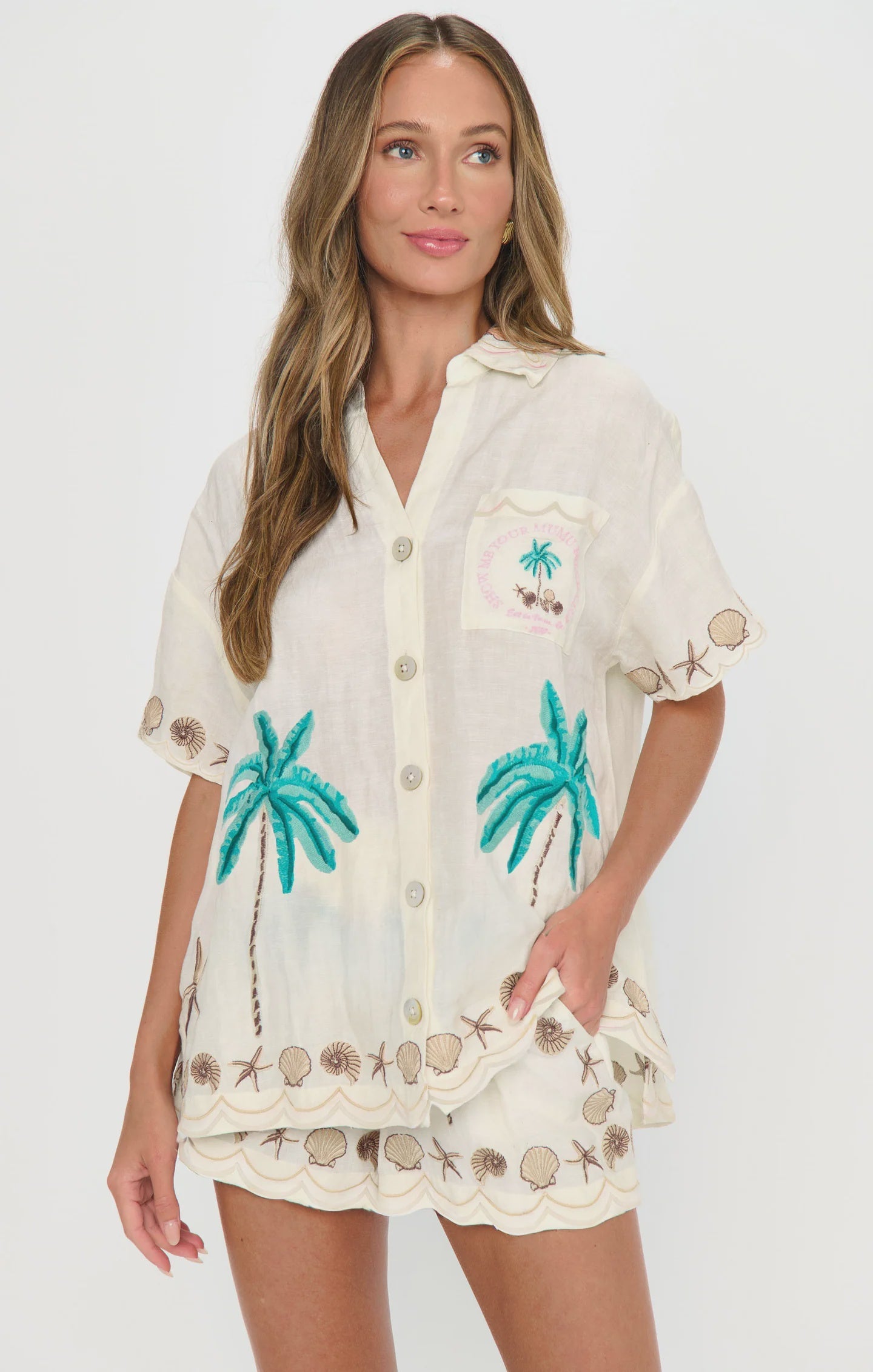 A woman in Zertz's HENDRIX SHORTS—cream-colored, high-waisted with embroidered palm trees and seashells—and a matching short-sleeve button-up shirt stands against a plain white background, her long wavy hair loose.