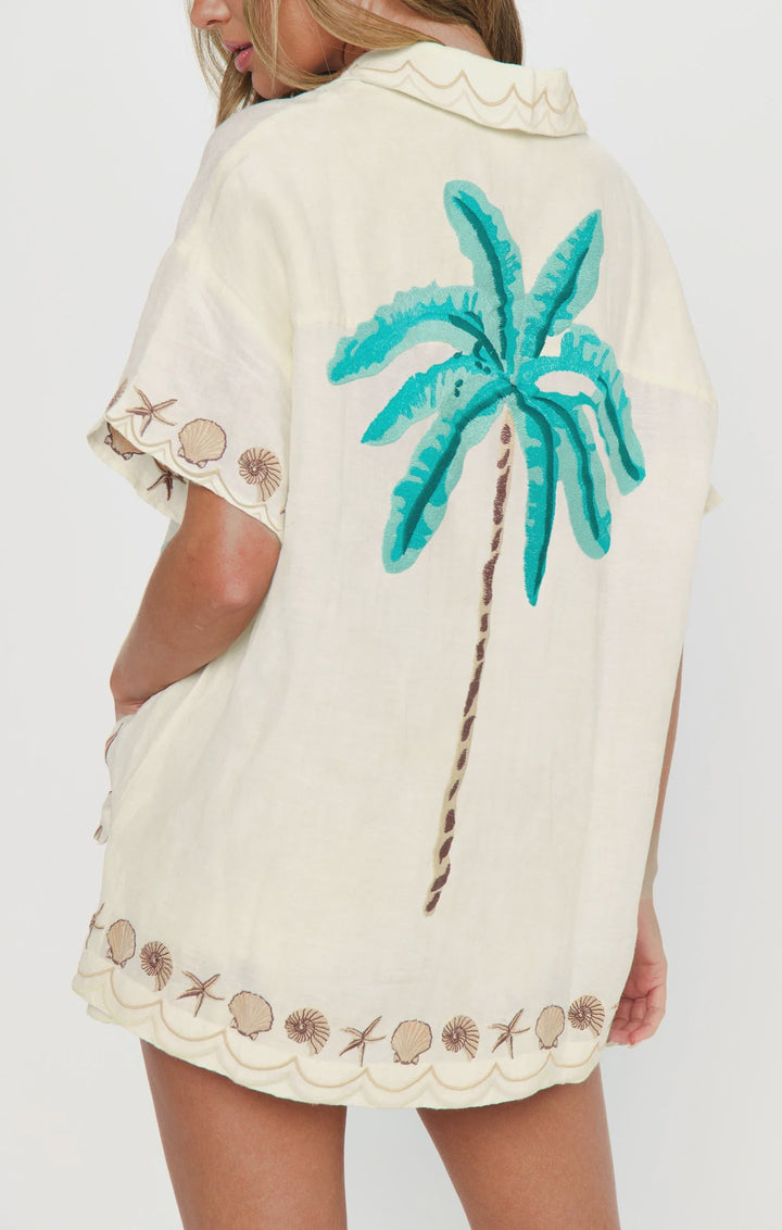 A person wearing the Zertz PARKER BUTTON DOWN TOP—an oversized cream shirt with brown seashell and wave embroidery at the hem and sleeves, and a large green palm tree embroidered on the back—is turned slightly away from the camera.
