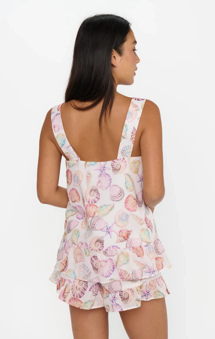 A woman with long dark hair is seen from the back wearing the LAYLA SET by Zertz— a matching tie-front top and ruffled shorts featuring a pastel seashell and starfish print, perfect for vacation, on a light background.