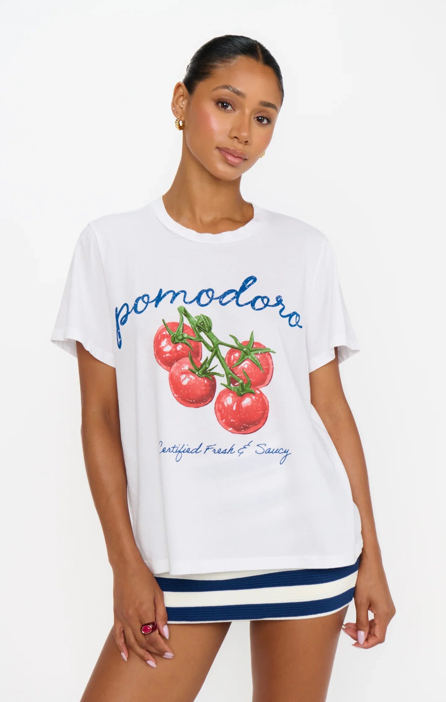 A woman poses against a white background wearing the Zertz TRAVIS TEE, an oversized fit top with illustrated tomatoes, styled with a blue and white striped mini skirt.