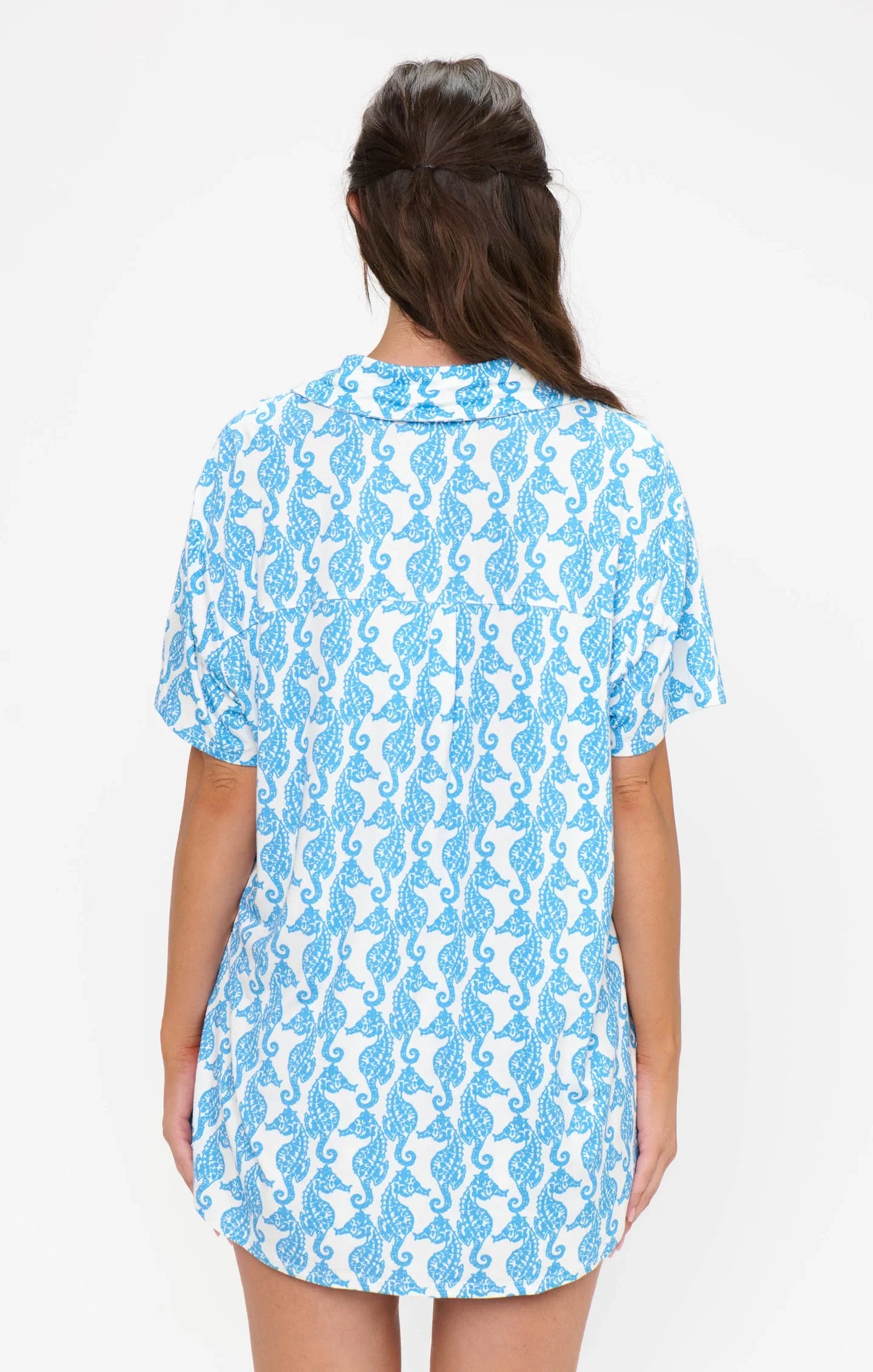A person with long brown hair stands facing away, wearing the Zertz SUNDAY MORNING PJ SET—a white short-sleeve button-down top and elastic waist shorts with a seahorse print—against a plain white background.