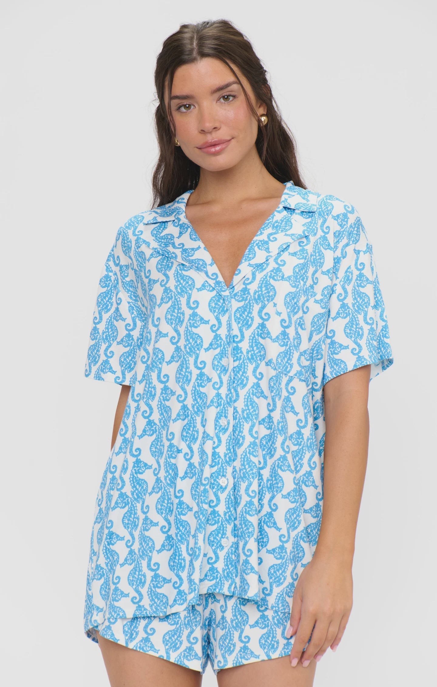 A woman wears the SUNDAY MORNING PJ SET by Zertz, featuring a blue and white seahorse print with a button-down top and elastic waist shorts, as she stands against a plain white backdrop.