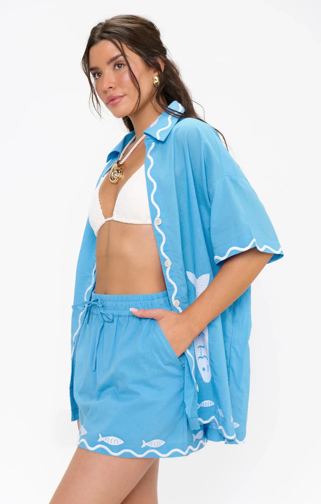 A woman with long brown hair wears the Zertz PARKER BUTTON DOWN over a white bikini top and fish-print shorts, accessorized with gold hoop earrings and a necklace, standing against a plain white background.