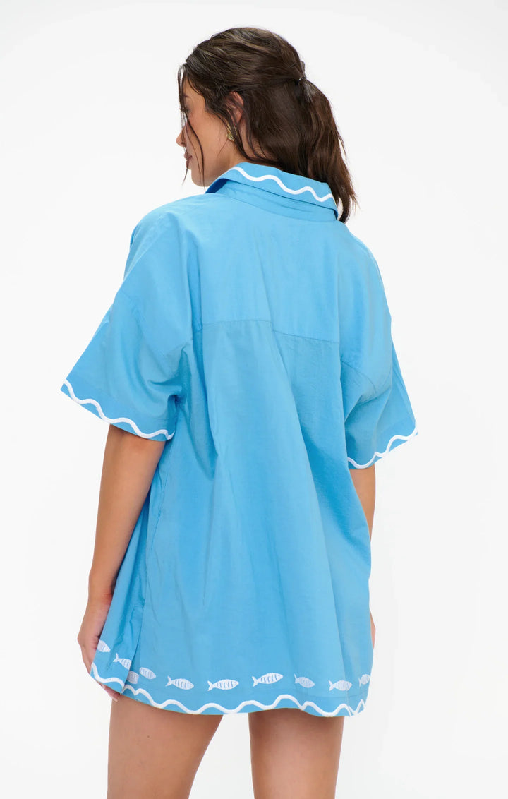 A woman with dark hair in a ponytail, seen from behind, is wearing the Zertz PARKER BUTTON DOWN—an oversized blue shirt with white wave and fish patterns along the edges.