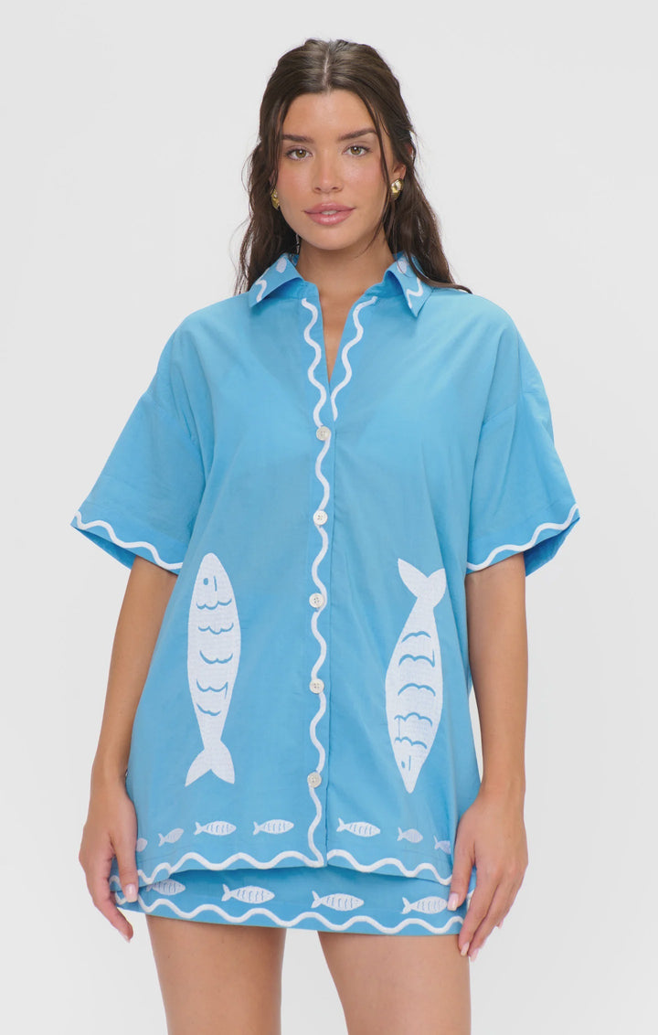 A woman models the Zertz PARKER BUTTON DOWN in light blue with white fish embroidery and wavy trim, standing against a plain white background.