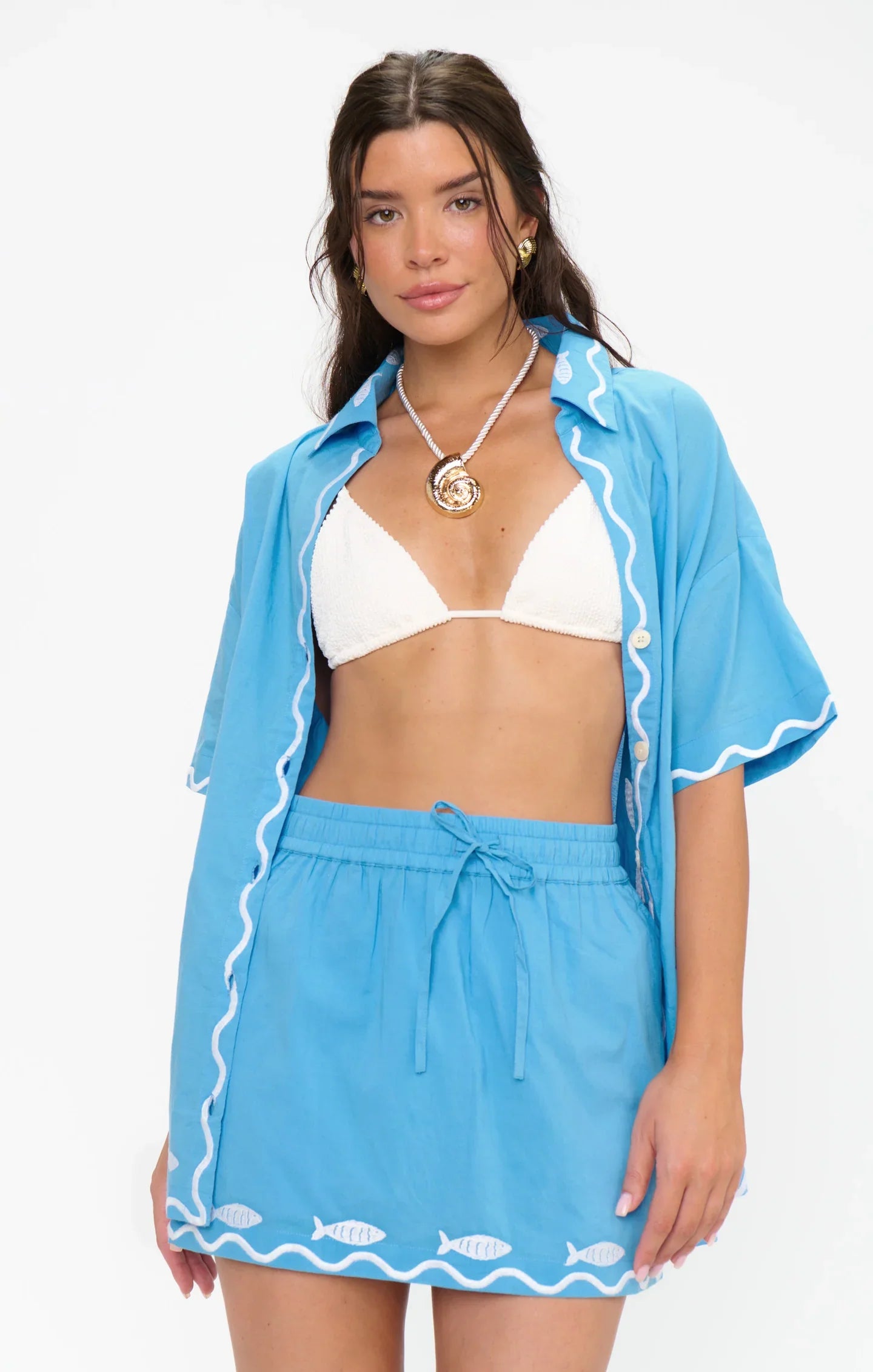 A woman wears a white bikini top under a loose blue shirt with white trim, paired with the BAY STREET SKIRT by Zertz—a blue skirt with a fish pattern and elastic drawstring waist—perfect for beachside adventures. She accessorizes with a necklace.