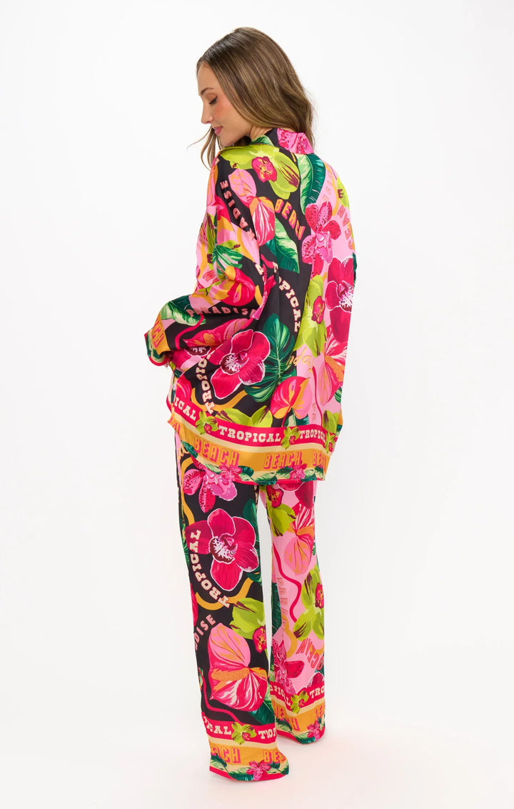 A person stands facing away, wearing Zertz’s HOT TROPIC NIGHTS PJ SET—a bold, colorful pantsuit with tropical floral prints in pink, yellow, green, and red. The vibrant pattern displays the words "Tropical" and "Fierce.