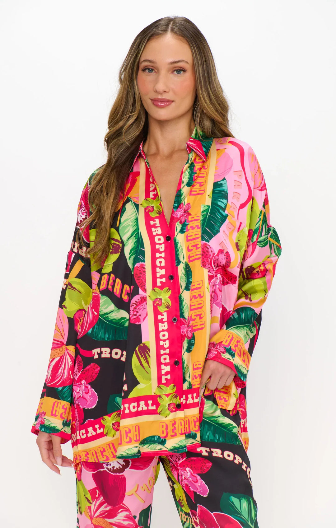 A woman with long, wavy hair wears the HOT TROPIC NIGHTS PJ SET by Zertz—a silky, oversized button-down and matching pants featuring a vibrant tropical print with words like TROPICAL and BEACH.