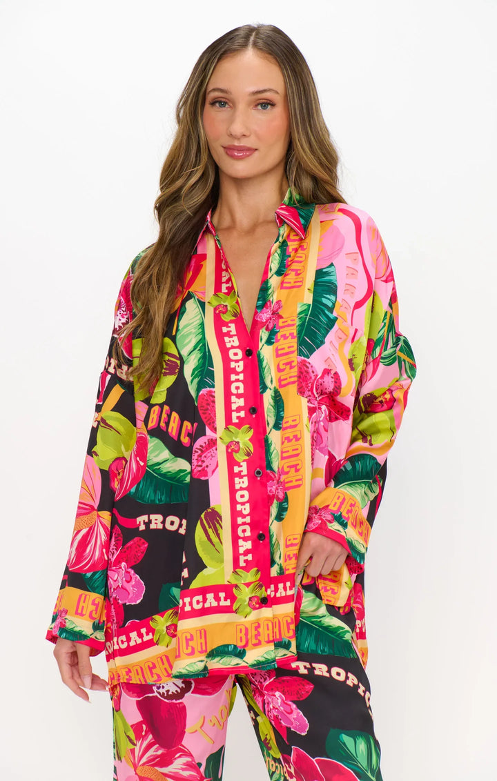 A woman with long, wavy hair wears the HOT TROPIC NIGHTS PJ SET by Zertz—a silky, oversized button-down and matching pants featuring a vibrant tropical print with words like TROPICAL and BEACH.