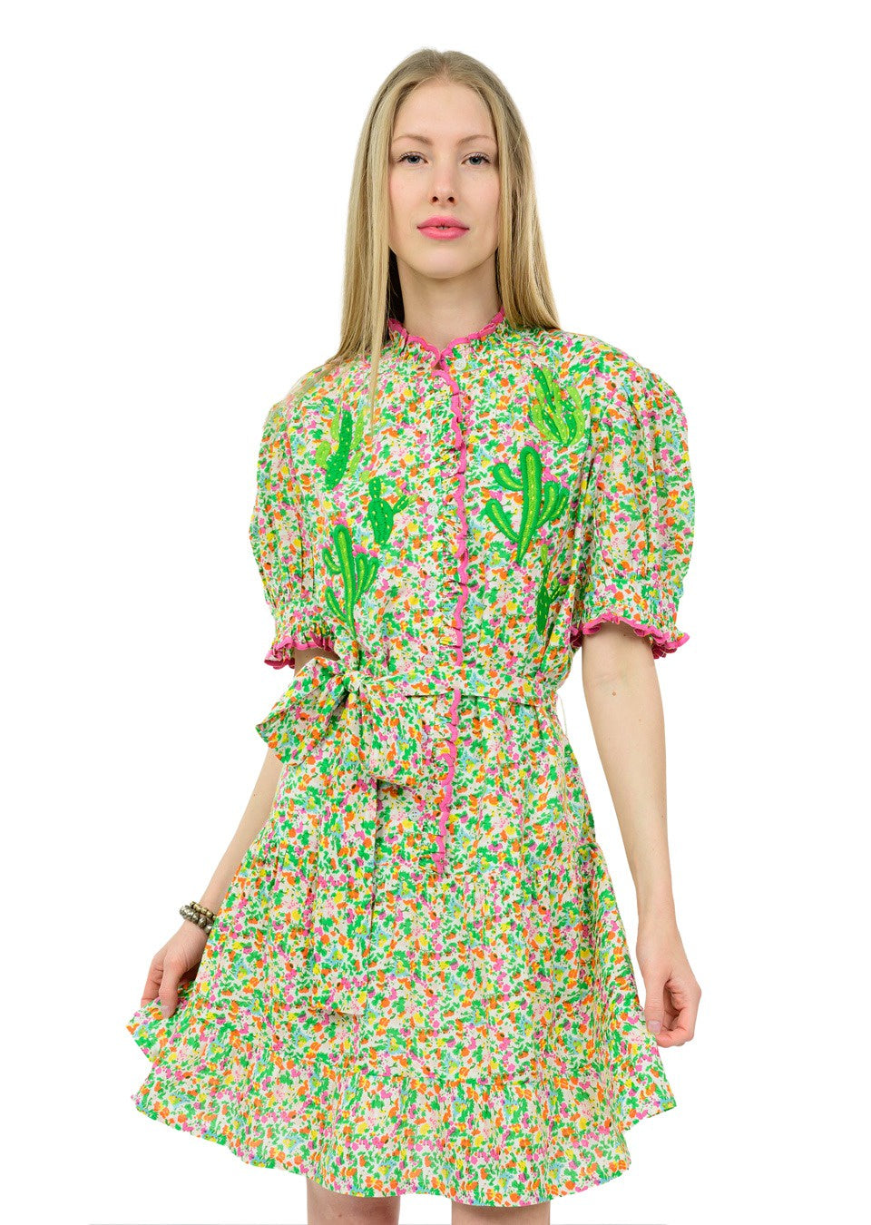 A woman models the Zertz NOPAL DRESS—featuring colorful floral prints, green cactus motifs, pink trim, short puff sleeves, and a tied waist—against a white background. Ideal for conscious fashion lovers.