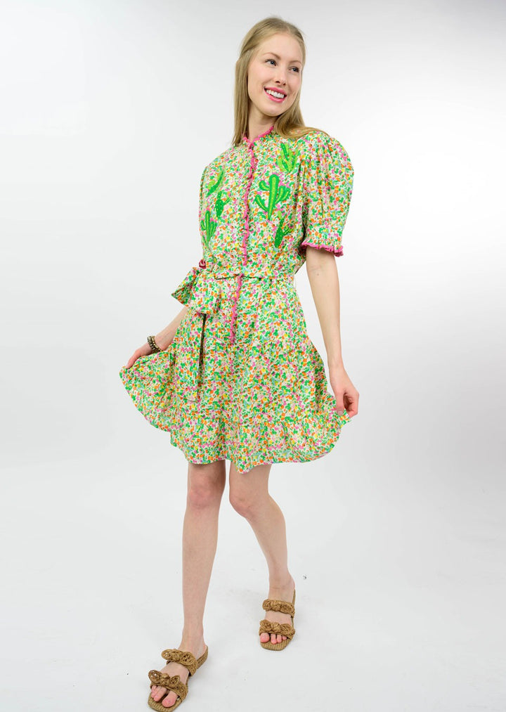 A woman smiles in the Zertz NOPAL DRESS, featuring colorful floral prints, puffy sleeves, and green leaf motifs. She poses with one hand holding her skirt and wears tan sandals, set against a plain white background.