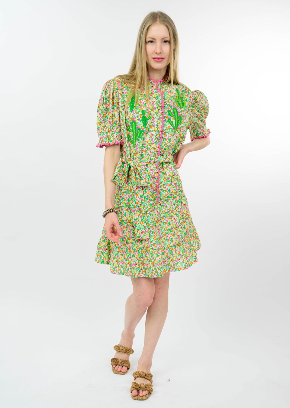 A woman with long blonde hair stands against a white background in the Zertz NOPAL DRESS—a vibrant, short-sleeve floral dress with green accents and ruffled edges—styled with tan sandals and a bracelet for a conscious fashion look.