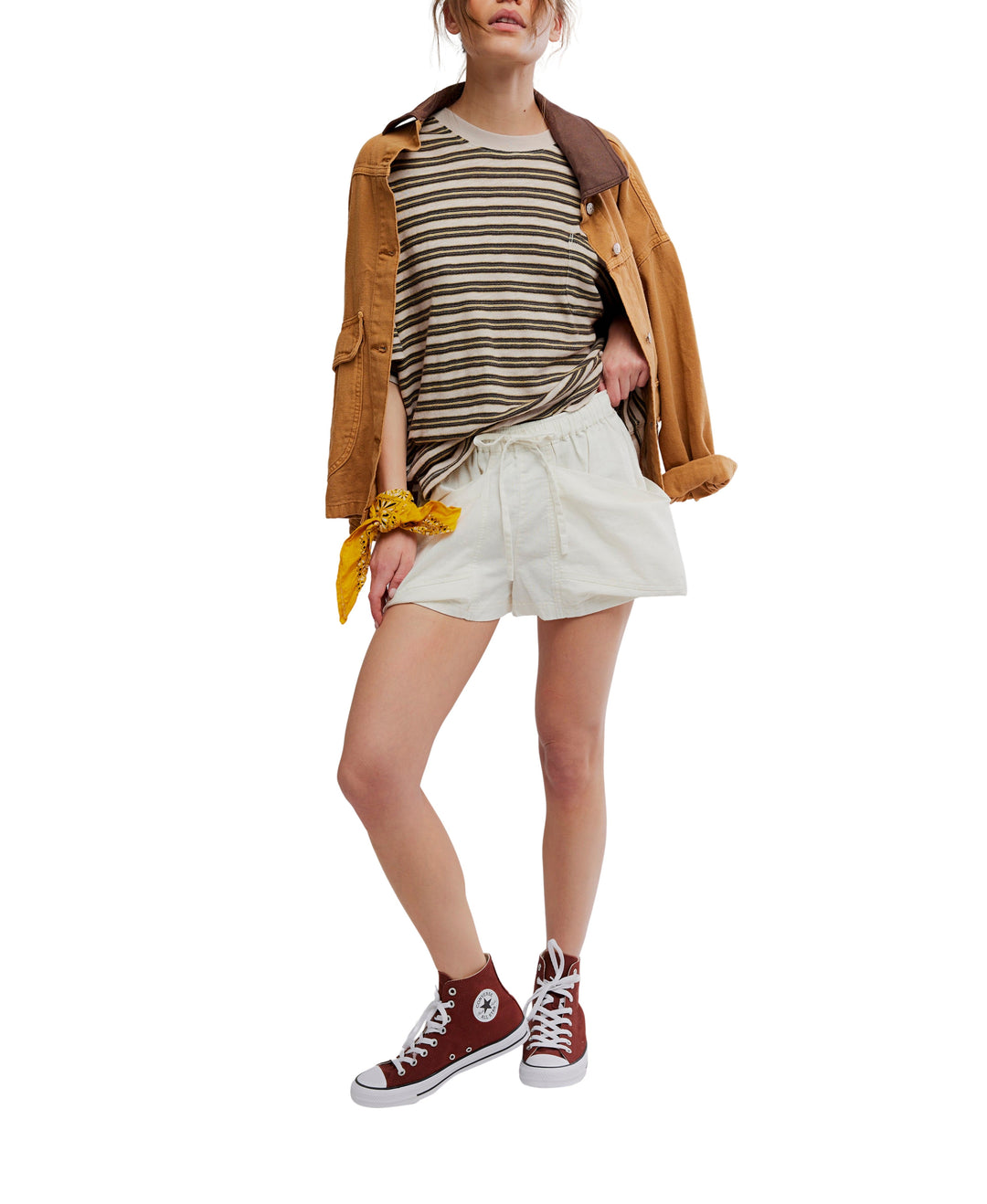A woman wears Zertz GOOD FORTUNE SHORTS, a striped tee, a brown jacket over her shoulders, maroon high-top sneakers, and a yellow bandana on her wrist. Her face is partially out of view.