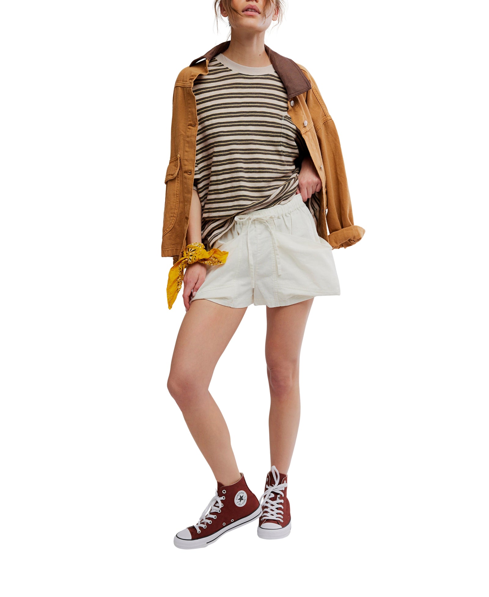 A woman wears Zertz GOOD FORTUNE SHORTS, a striped tee, a brown jacket over her shoulders, maroon high-top sneakers, and a yellow bandana on her wrist. Her face is partially out of view.