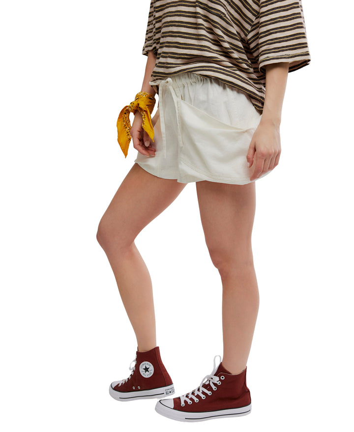 Against a white background, a person wears a striped brown and white t-shirt, Zertz GOOD FORTUNE SHORT cream-colored shorts, red high-top Converse sneakers, and a yellow bandana tied around their wrist.