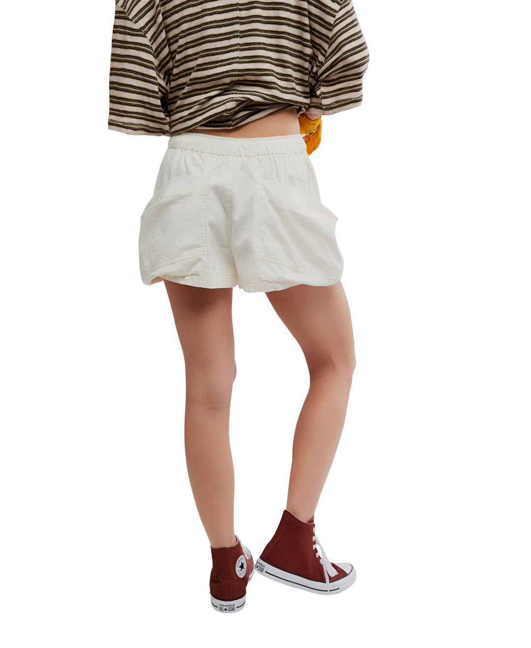 A person stands with one leg bent, facing away in a brown and white striped shirt, Zertz GOOD FORTUNE SHORTS in white, and maroon high-top sneakers against a white background.