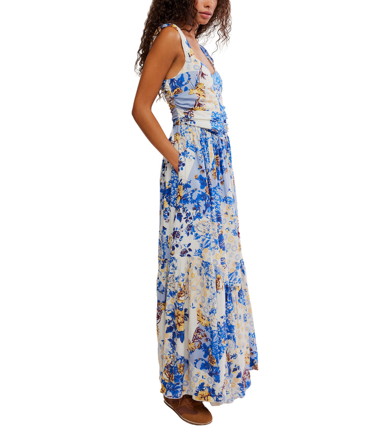 A woman stands sideways in the SASHA MAXI dress by Zertz—a blue maxi with white botanical print and pockets. She has curly hair, brown flat shoes, and is set against a white background that highlights her elegant look.