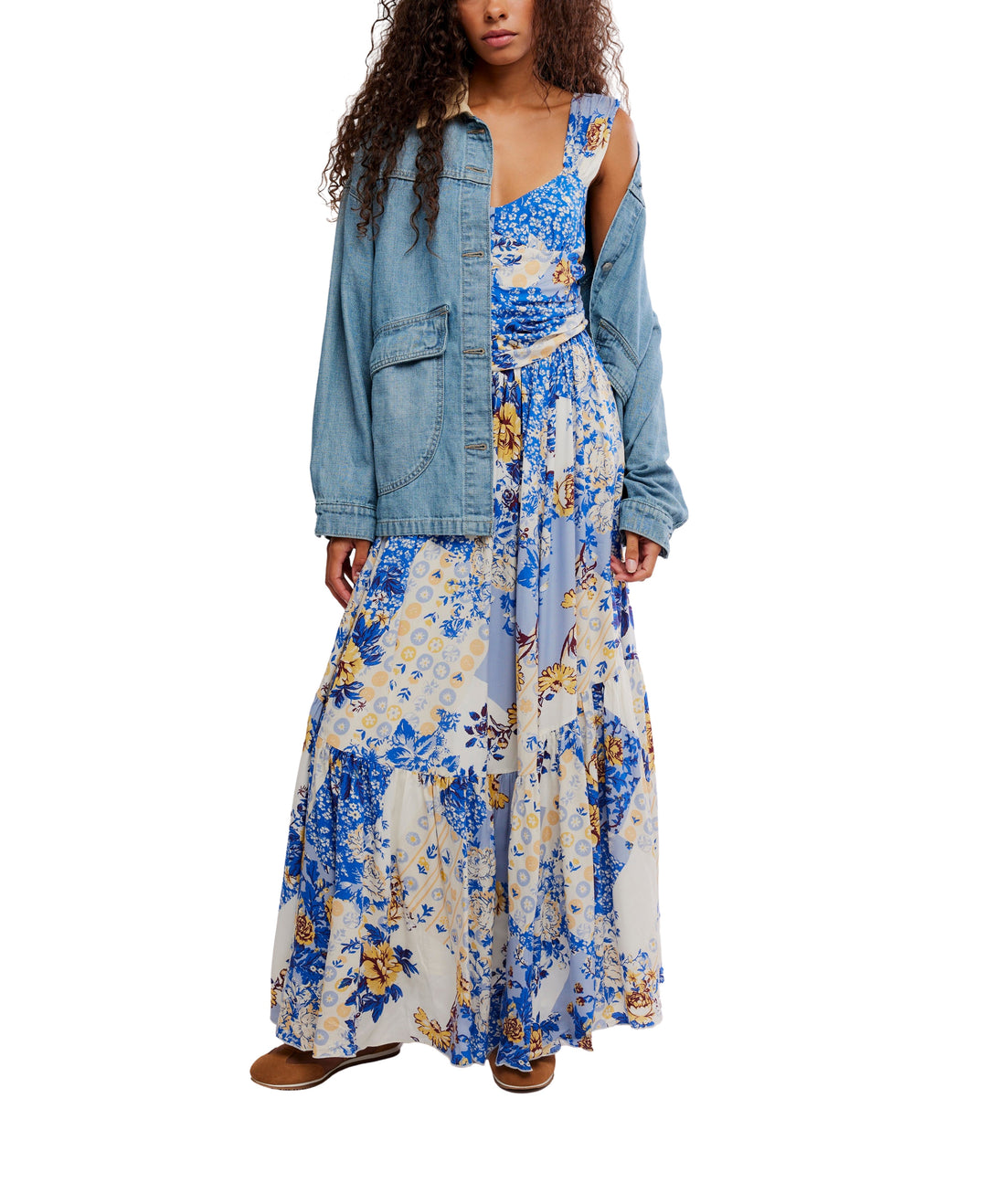 A person wearing the SASHA MAXI blue floral patchwork dress by Zertz and a loose denim jacket draped over one shoulder, paired with brown shoes, stands against a white background.