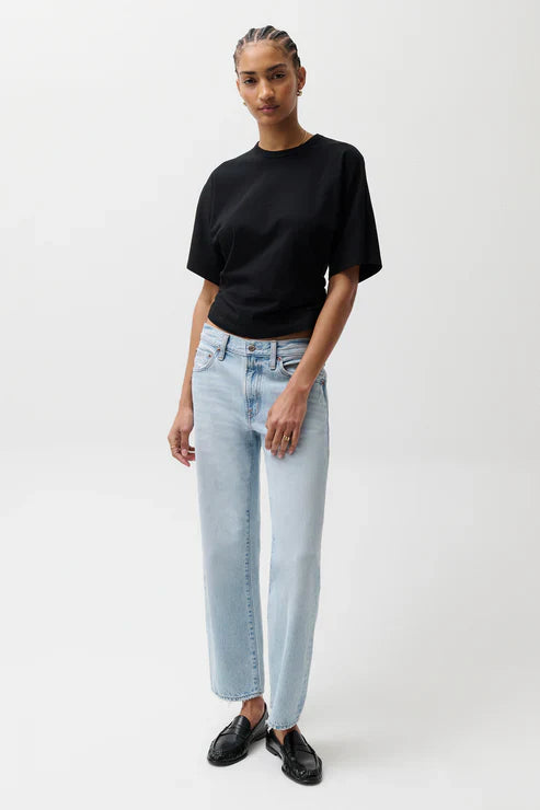 A person stands against a white background wearing a black t-shirt, black loafers, and Zertz LEXI MID RISE BOWED STRAIGHT jeans with a vintage wash. They have braided hair and a relaxed expression, with one hand by their side.
