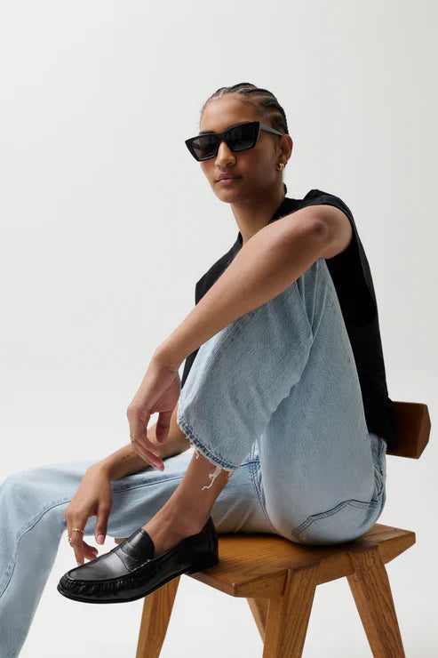 A person wearing black sunglasses, a black T-shirt, Zertz LEXI MID RISE BOWED STRAIGHT jeans, and black loafers sits sideways on a wooden stool against a plain white background.