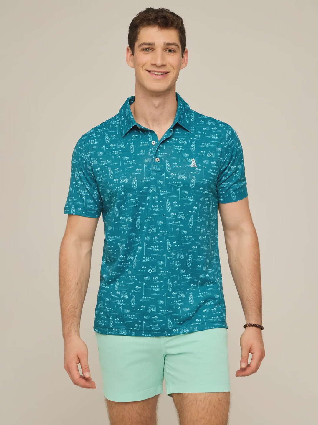 A man stands smiling in a teal Zertz THE FAIRWAGER PERFORMANCE POLO with a white pattern, paired with light green shorts. The polo is crafted from performance knit fabric. The background is plain and neutral.