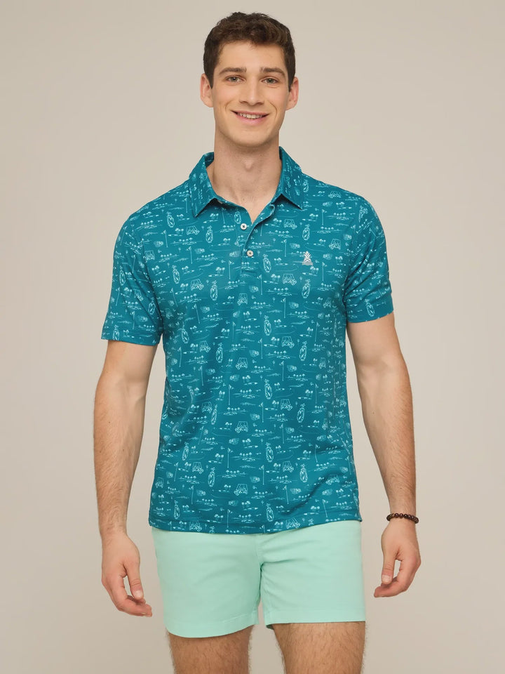 A man stands smiling in a teal Zertz THE FAIRWAGER PERFORMANCE POLO with a white pattern, paired with light green shorts. The polo is crafted from performance knit fabric. The background is plain and neutral.