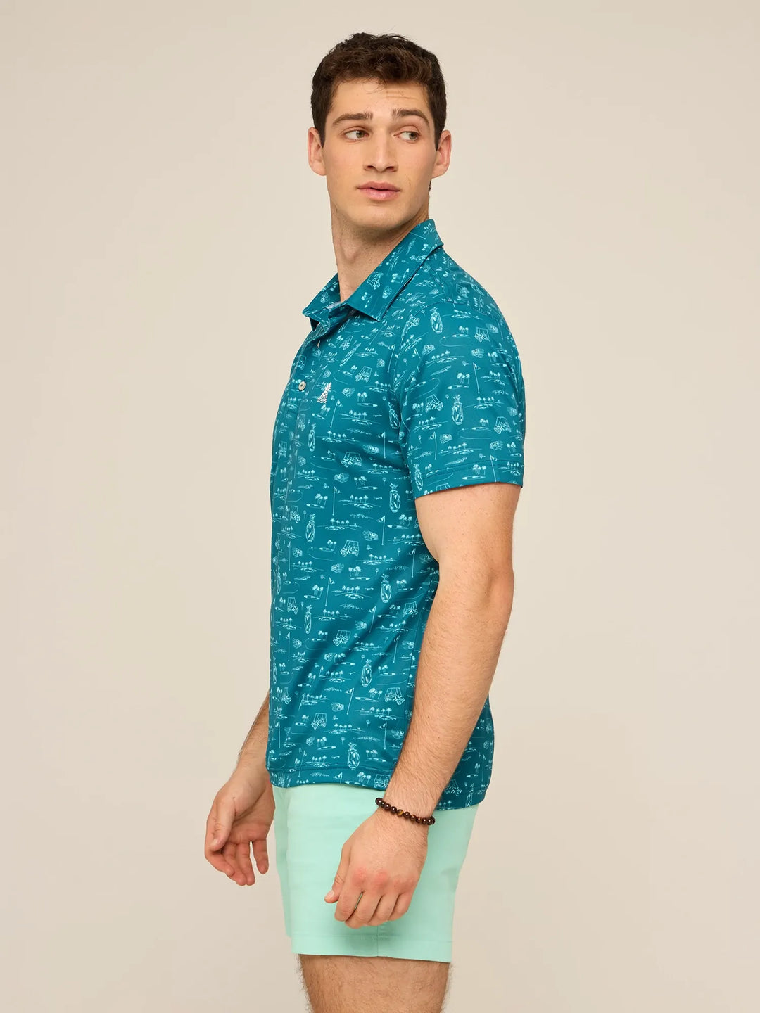 A man stands sideways in a teal Zertz THE FAIRWAGER PERFORMANCE POLO with a patterned design and light aqua shorts, looking over his shoulder against a plain beige background.
