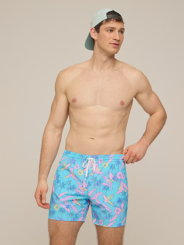 A man wearing a backward light green cap and Zertz THE FLOCK PARTIES 5.5" CLASSIC LINED SWIM TRUNK with a pink tropical pattern stands shirtless against a plain background, looking to the side with one hand on his hip.