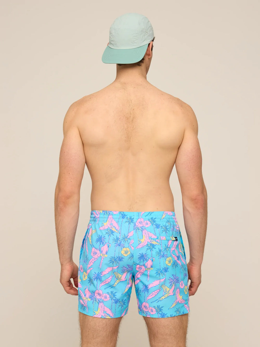 A man seen from behind wearing a light blue cap and Zertz THE FLOCK PARTIES 5.5" CLASSIC LINED SWIM TRUNK, featuring a vibrant tropical print with pink flamingos and green palm leaves, stands against a plain beige background.