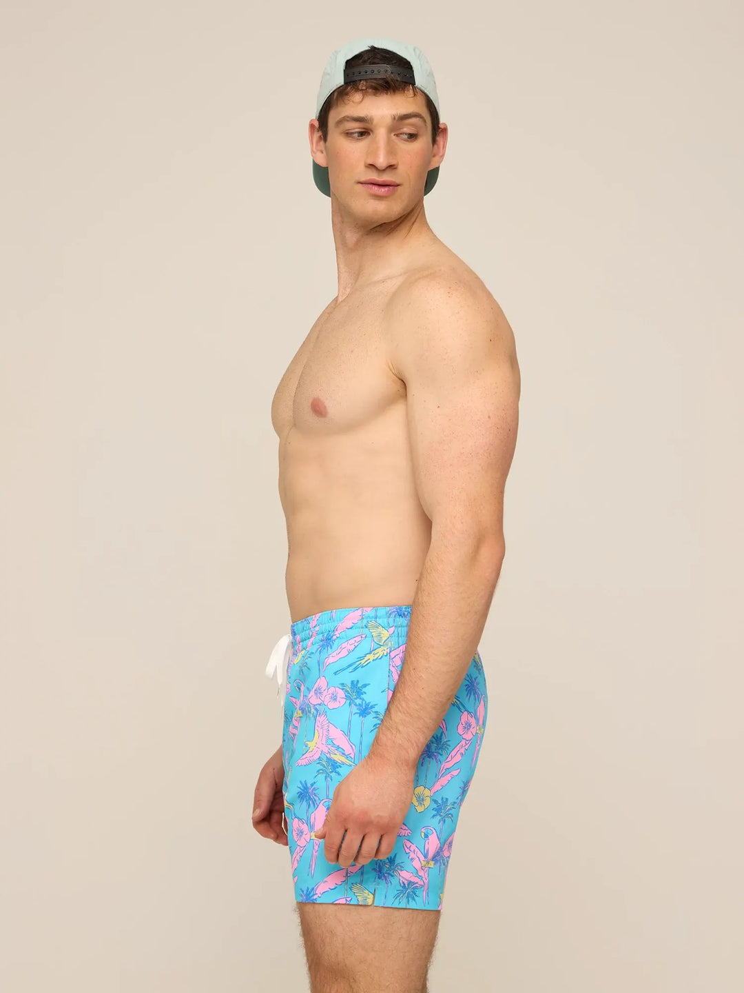 A man in a light blue cap and Zertz THE FLOCK PARTIES 5.5" CLASSIC LINED SWIM TRUNK, featuring a vibrant tropical pattern, stands sideways and looks over his shoulder against a plain beige background.