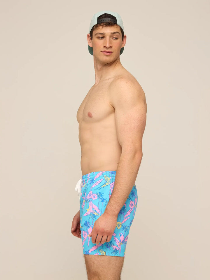 A man in a light blue cap and Zertz THE FLOCK PARTIES 5.5" CLASSIC LINED SWIM TRUNK, featuring a vibrant tropical pattern, stands sideways and looks over his shoulder against a plain beige background.