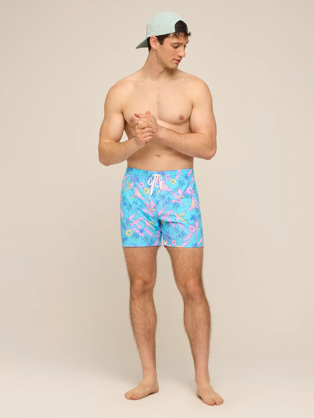 A shirtless man in Zertz THE FLOCK PARTIES 5.5" CLASSIC LINED SWIM TRUNK and a light blue cap stands barefoot against a beige background, looking left with his hands clasped.