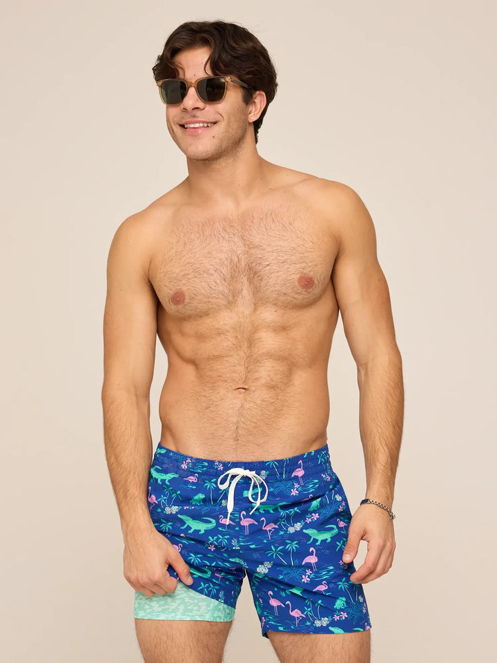 A smiling man with brown hair and sunglasses models Zertz THE SWAMP &amp; CIRCUMSTANCES 5.5" Classic Lined Swim Trunks, featuring a quick-dry tropical flamingo and alligator print, against a plain beige background.