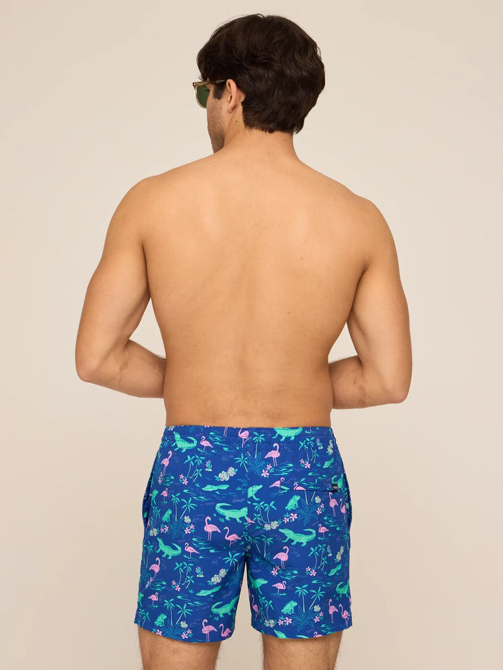 A man faces away from the camera, shirtless, wearing Zertz THE SWAMP &amp; CIRCUMSTANCES 5.5" Classic Lined Swim Trunks with a tropical flamingo, palm, and green foliage print on a light beige background.