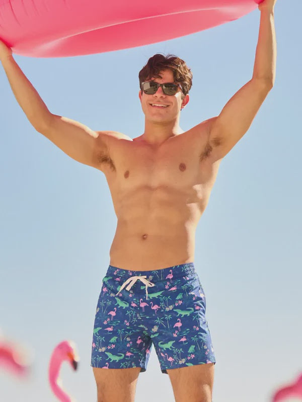 A smiling man wears Zertz THE SWAMP &amp; CIRCUMSTANCES 5.5" CLASSIC LINED SWIM TRUNKS featuring a blue flamingo pattern while holding a large pink inflatable, with clear blue sky and plastic pink flamingos visible below.