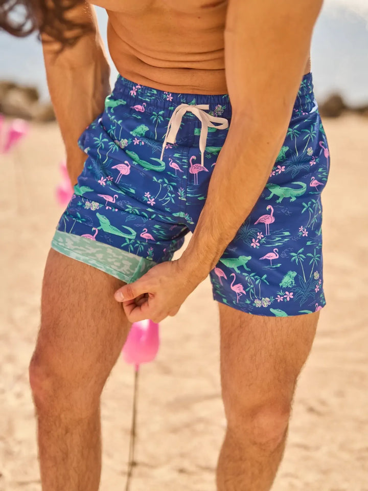 On a sandy beach, someone pulls up the leg of their Zertz THE SWAMP &amp; CIRCUMSTANCES 5.5" CLASSIC LINED SWIM TRUNKS, featuring playful green alligators, pink flamingos, and palm trees; pink flamingo lawn ornaments appear in the background.