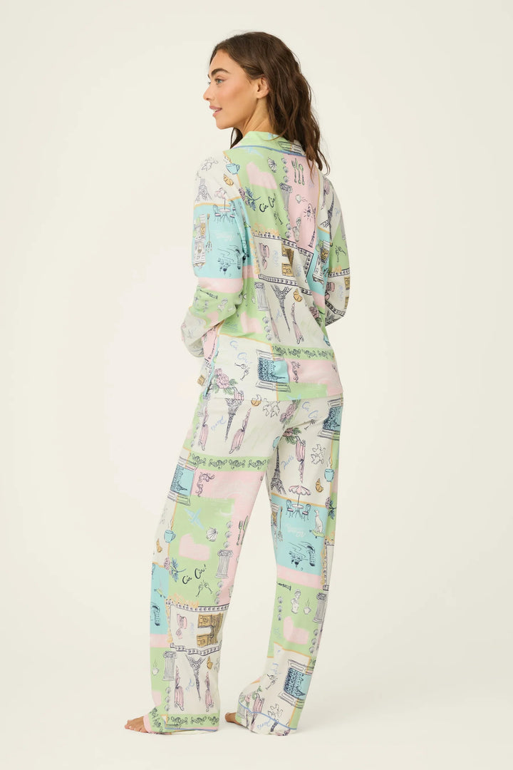 A woman faces away, wearing the Zertz WHIMSY POSTCARDS PJ SET—a cozy, loose-fitting, long-sleeved cotton-modal loungewear set in pastel pink, green, blue, and cream with a whimsical illustrated pattern.