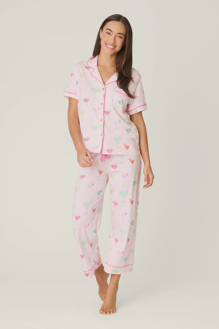A woman smiles barefoot in the Zertz WHIMSY SIP HAPPENS PJ SET—light pink women’s pajamas with a cocktail print, featuring a short-sleeve button-up top and long pants—her hair down against a plain background.