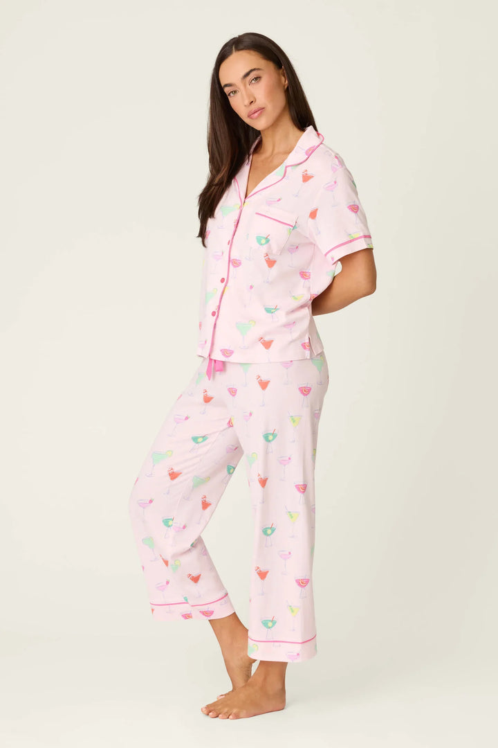 A woman stands barefoot in the Zertz WHIMSY SIP HAPPENS PJ SET—light pink, short-sleeved with pink piping. This comfy set features button closure, a chest pocket, and playful cocktail prints on an off-white background.