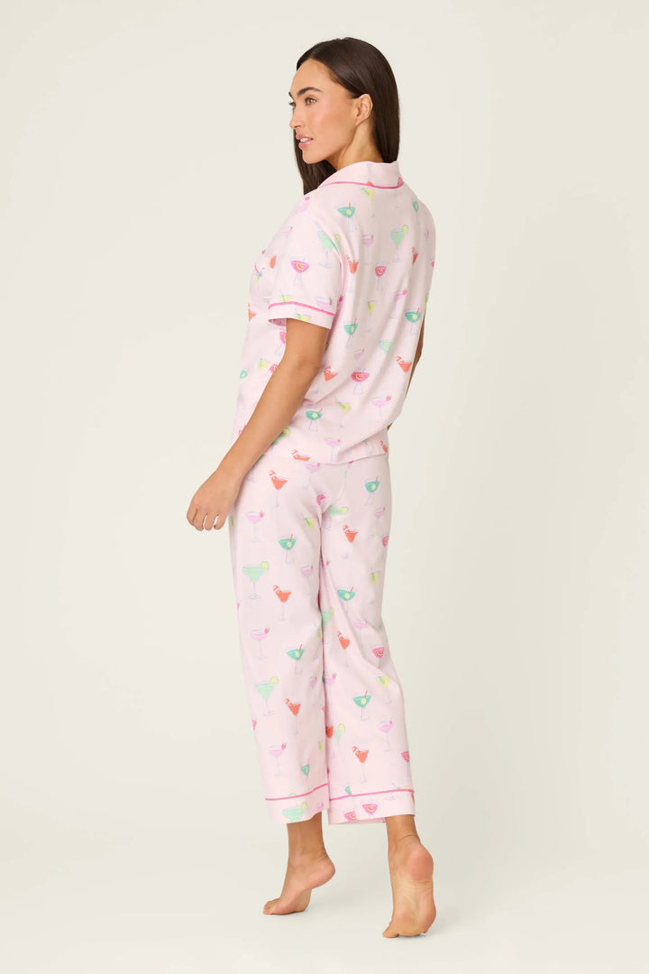 A woman stands barefoot on a light background, wearing the Zertz WHIMSY SIP HAPPENS PJ SET—a light pink, short-sleeve button-up and pants featuring a colorful cocktail glass pattern. She looks over her shoulder.