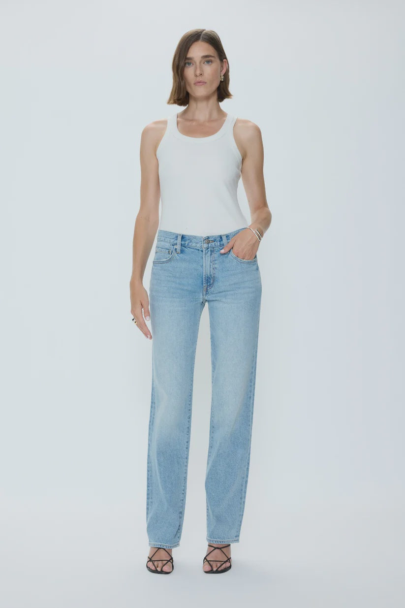 A woman poses against a white background, wearing the Zertz ISSA MID RISE RELAXED STRAIGHT jeans in light blue rigid denim with a white sleeveless top, black strappy heels, minimal jewelry, and a sleek bob hairstyle.