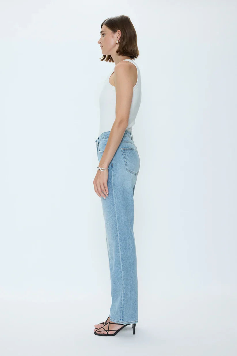 A woman stands in profile against a plain white background, wearing a white sleeveless top, Zertz ISSA MID RISE RELAXED STRAIGHT jeans, black heeled sandals, and simple jewelry.