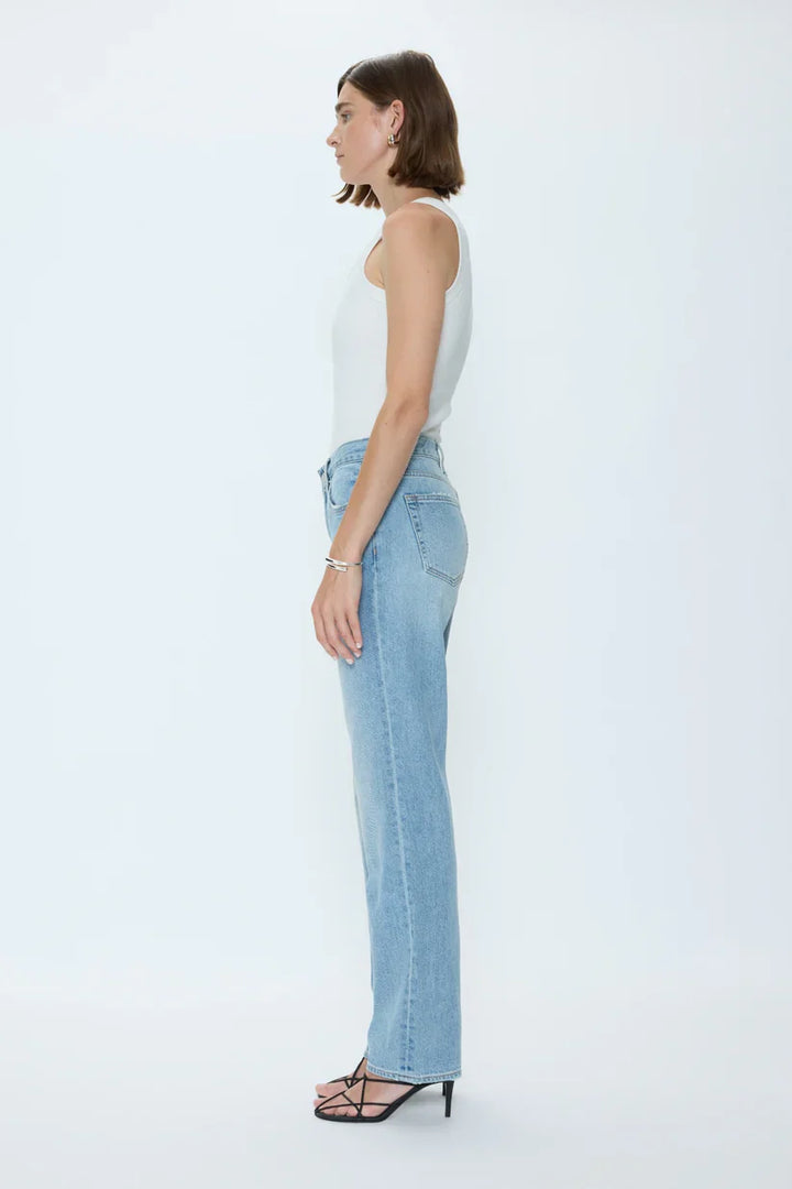 A woman stands in profile against a plain white background, wearing a white sleeveless top, Zertz ISSA MID RISE RELAXED STRAIGHT jeans, black heeled sandals, and simple jewelry.