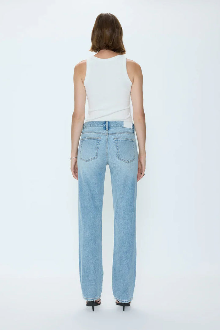 A person with shoulder-length brown hair, in a white sleeveless top, Zertz ISSA MID RISE RELAXED STRAIGHT jeans, and black heeled sandals stands facing away against a plain white background.